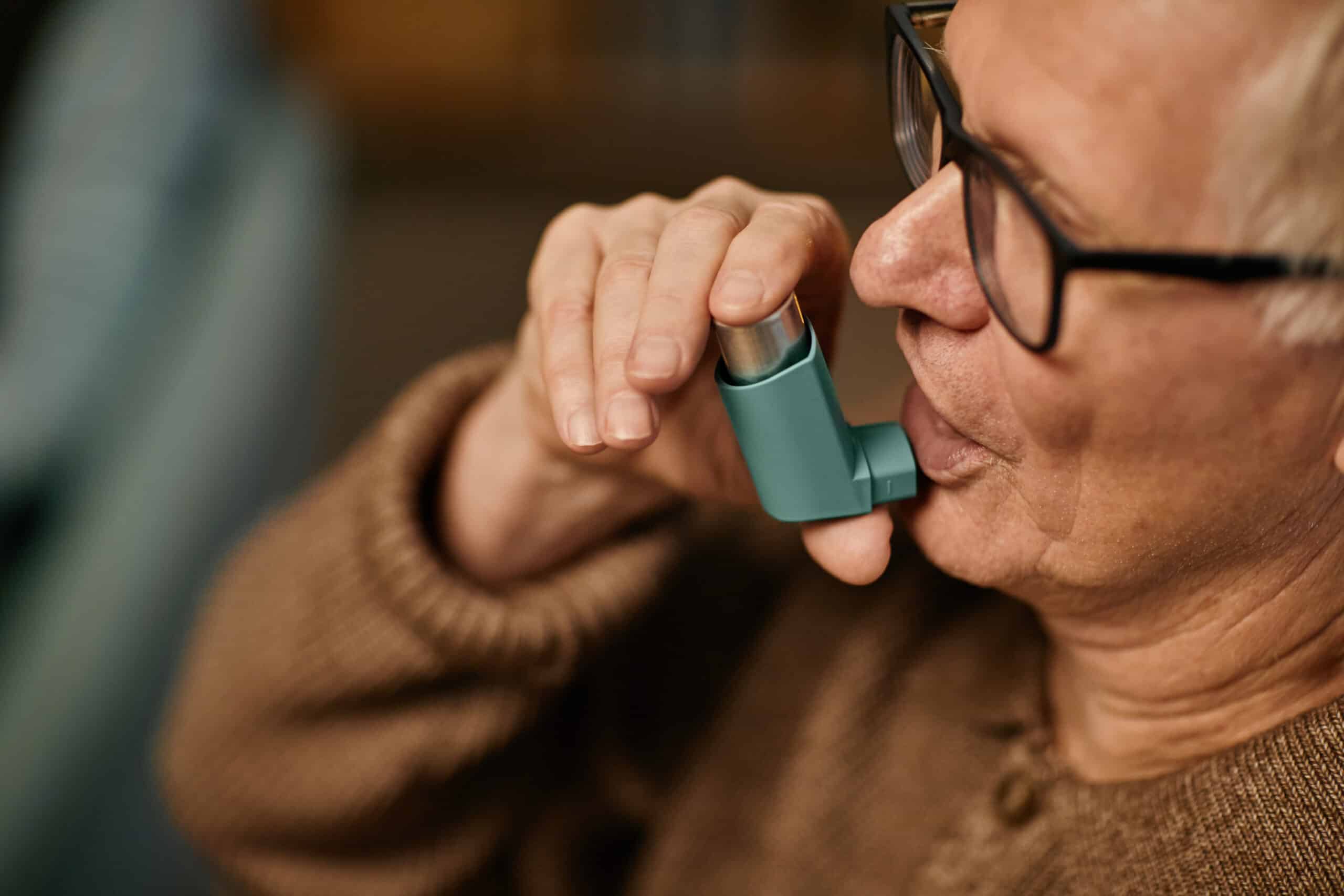 Senior Caucasian man using inhaler while sitting in nursing home, holding device close to mouth, wearing eyeglasses, focusing on managing respiratory condition, partial side view asthma attack